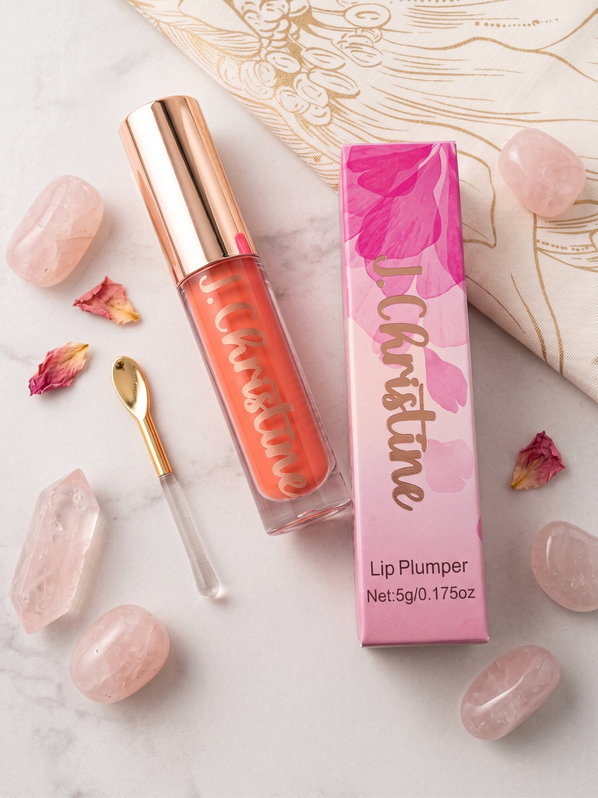 A liquid lip plumper in a clear tube with a metallic rose gold cap, shown next to its pink and white packaging box. The product is styled on a white marble surface with rose quartz crystals, dried rose petals, and a gold spoon.